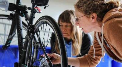 Two women examine the wheel of a bicycle that is being repaired indoors