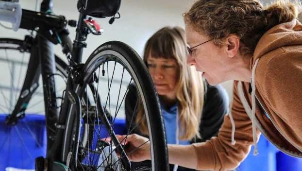 Two women examine the wheel of a bicycle that is being repaired indoors