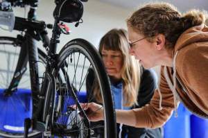 Two women examine the wheel of a bicycle that is being repaired indoors