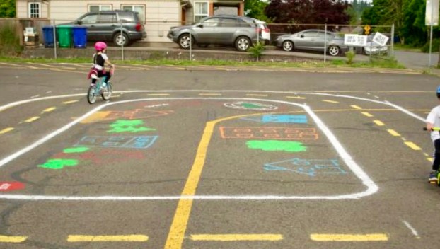 Two kids wearing helmets bike along marked lines on pavement that mimic street roads and signs