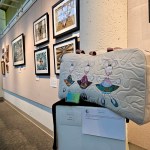 A GALA art exhibit inside Gresham City Council Chambers featuring an ornate bag on a pedestal in front of a wall of paintings